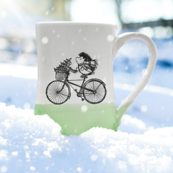Handmade coffee mug with drawing of hedgehog delivering a Christmas tree on a bike. Green accent color.