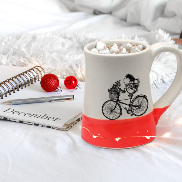Handmade coffee mug with drawing of hedgehog delivering a Christmas tree on a bike. Red accent color.