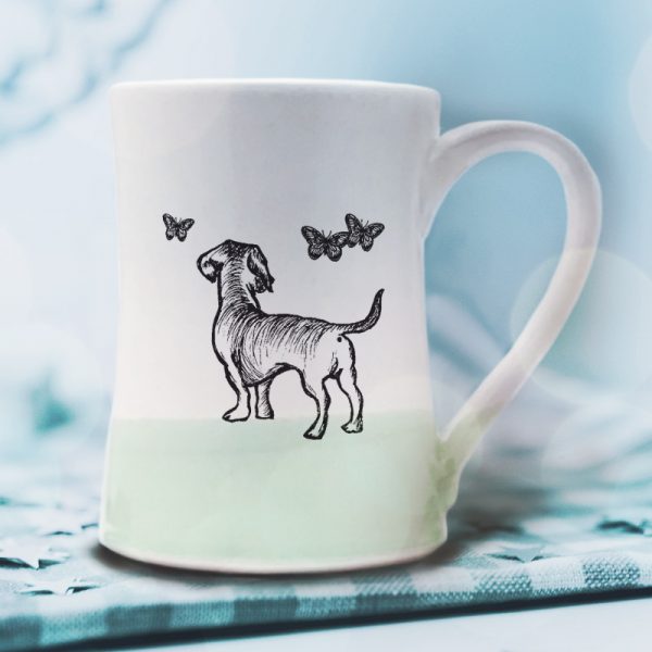 A handmade coffee mug with a drawing of a doggie hanging out with a bunch of butterflies. Green accent color.