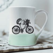 A handmade coffee mug with a drawing of hedgehog on a bike delivering a basket of flowers. Green accent color.