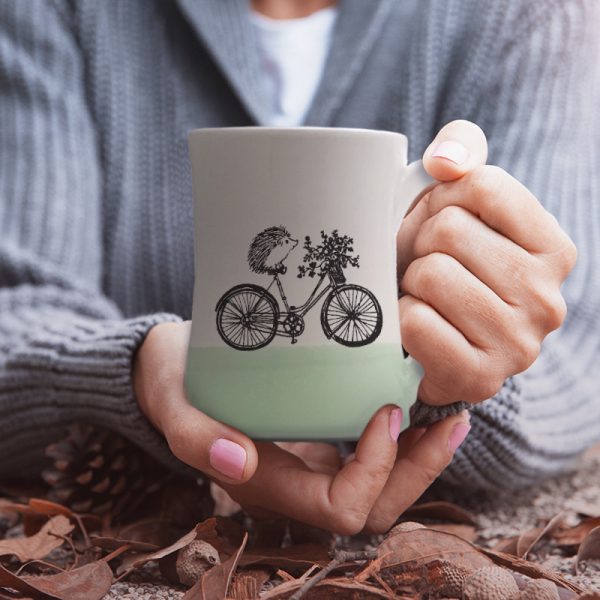 A handmade coffee mug with a drawing of hedgehog on a bike delivering a basket of flowers. Green accent color.