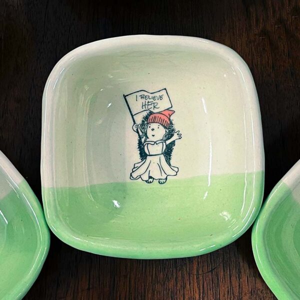 closeup of a handmade dish with a drawing of a hedgehog holding a sign that says I believe her