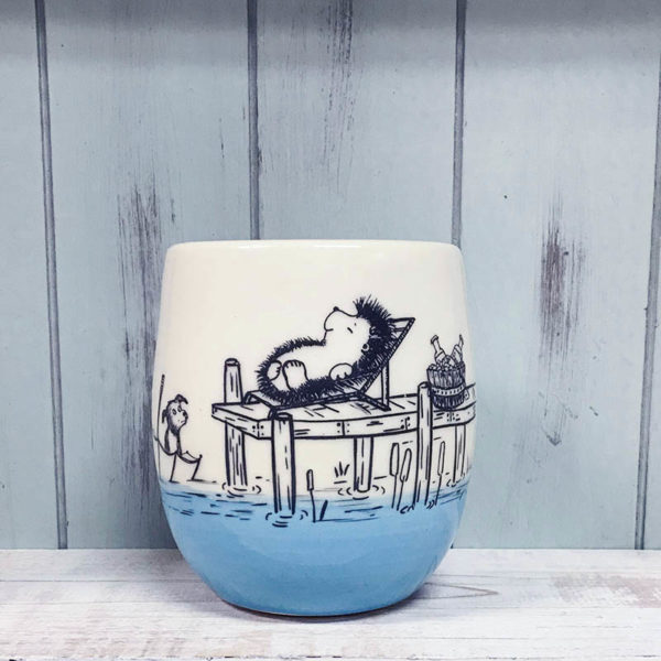 egg shaped cup with drawing of a hedgehog lounging on a dock while a dog swims around in an overturned umbrella