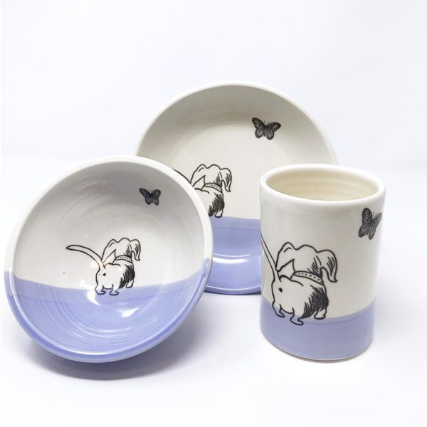 Collection of handmade pottery with a drawing of a dashchund chasing a butterfly. Grouping contains bowl, plate and tumbler with lavender accent. Made by hedgehogs