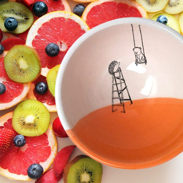 A handmade ceramic soup bowl with a drawing of a little bear in a swing and a hedgehog on a ladder. Coral accent color.