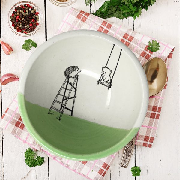 A handmade ceramic soup bowl with a drawing of a little bear in a swing and a hedgehog on a ladder. Green accent color.