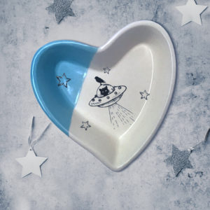 Handmade ceramic heart shaped dish with a drawing of a cat in a flying saucer or UFO. Blue accent color.