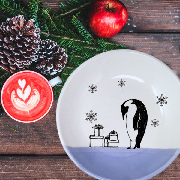 Handmade ceramic plate with a drawing of an Emperor Penguin examining his stack of presents in the snow. Lavender accent color.