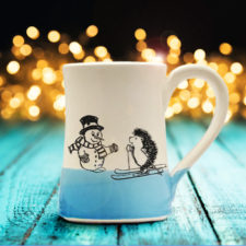 Handmade coffee mug with a drawing of a hedgehog on skis greeting a well-dressed snowman. Blue accent color