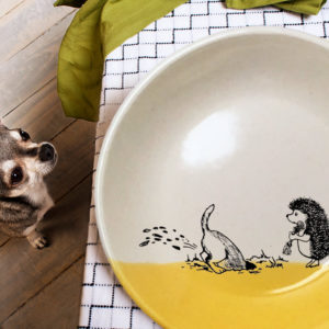 This ceramic plate is handmade by hedgehogs at Darn Pottery and has a drawing oof a dog digging in the yard while hedgehog looks on. Gold accent color
