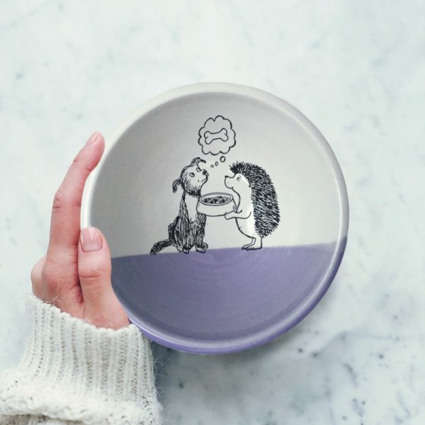 This handmade ceramic soup bowl has a cute drawing of a hedgehog offering a bowl of kibble to a dog. The dog, meanwhile, is dreaming of a bone. Lavender accent color