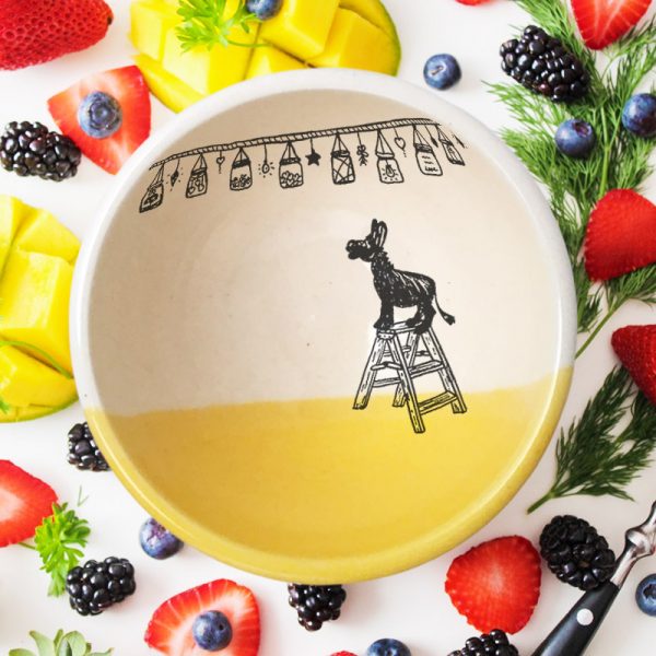 This soup or cereal bowl is handmade and features a drawing of a tiny donkey, standing on a step-ladder, looking at a string of lights overhead. Gold accent color.