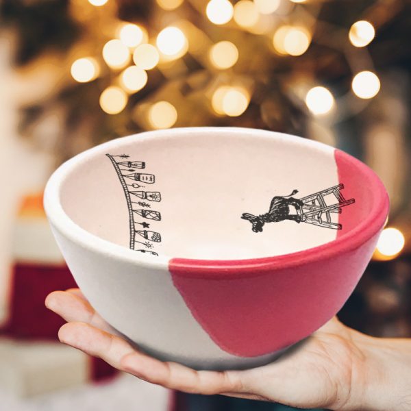This soup or cereal bowl is handmade and features a drawing of a tiny donkey, standing on a step-ladder, looking at a string of lights overhead. Red accent color.