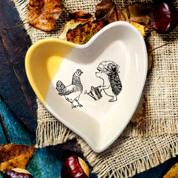 Handmade ceramic heart-shaped dish with drawing of a famer hedgehog talking to her chicken. Gold accent color