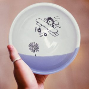 A hedgehog flying a biplane over the treetops. This is the original illustration on this handmade certamic soup bowl. Lavender accent color