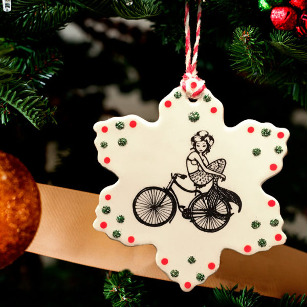 Handmade ceramic ornament with drawing of a mermaid riding a bicycle. Reverse side text reads Nevertheless, she persisted.