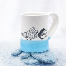 Handmade coffee mug with drawing of hedgehog pulling a Christmas tree on a little wagon. Blue accent color.