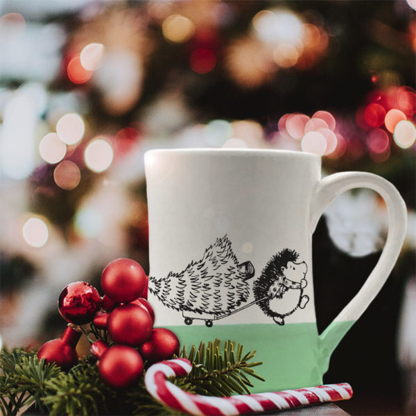 Handmade coffee mug with drawing of hedgehog pulling a Christmas tree on a little wagon. Green accent color.