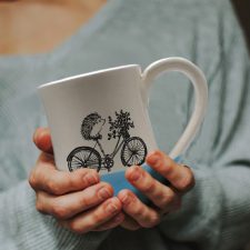 A handmade coffee mug with a drawing of hedgehog on a bike delivering a basket of flowers. Blue accent color.