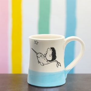 Handmade coffee mug with original illustration of a hedgehog riding on the back of a narwhal. Light blue accent color. Made by little crafty hedgehogs.