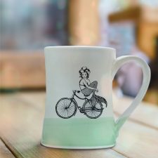 Handmade coffee mug with an undeterred mermaid riding a bicycle. She has a tail, nevertheless she is persisting. Green accent color.