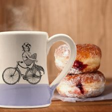 Handmade coffee mug with an undeterred mermaid riding a bicycle. She has a tail, nevertheless she is persisting. Lavender accent color.