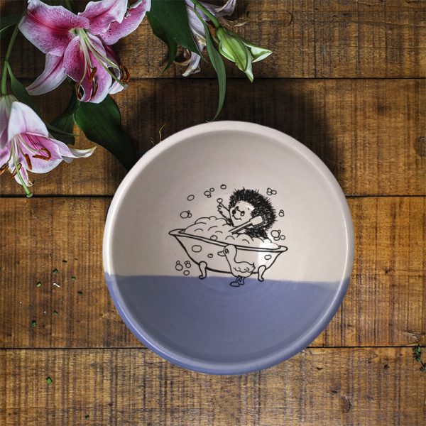This handmade clay bowl features a funny drawing of a hedgehog taking a bubble bath in a claw-footed bathtub. Lavender accent color.