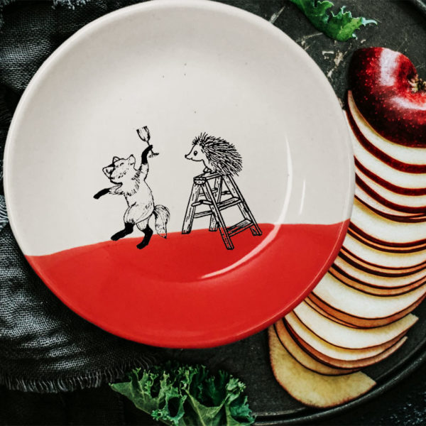 This plate is handmade by the hedgehogs of Darn Pottery and has a drawing of a fox celebrating with a champagne and a hedgehog climbing a ladder. Red accent color.