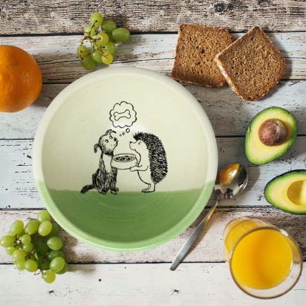This handmade ceramic soup bowl has a cute drawing of a hedgehog offering a bowl of kibble to a dog. The dog, meanwhile, is dreaming of a bone. Green accent color