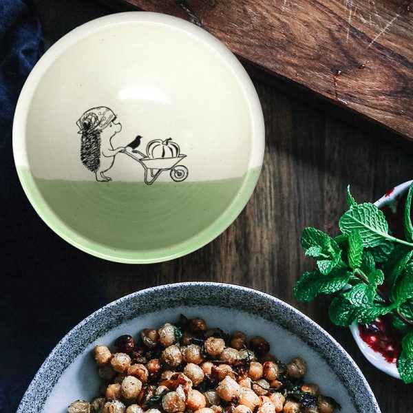 A happy little hedgehog is rolling a wheelbarrow with a giant pumpkin. This cute illustration is featured on a lovely handmade ceramic soup or salad bowl from Darn Pottery. Green accent color