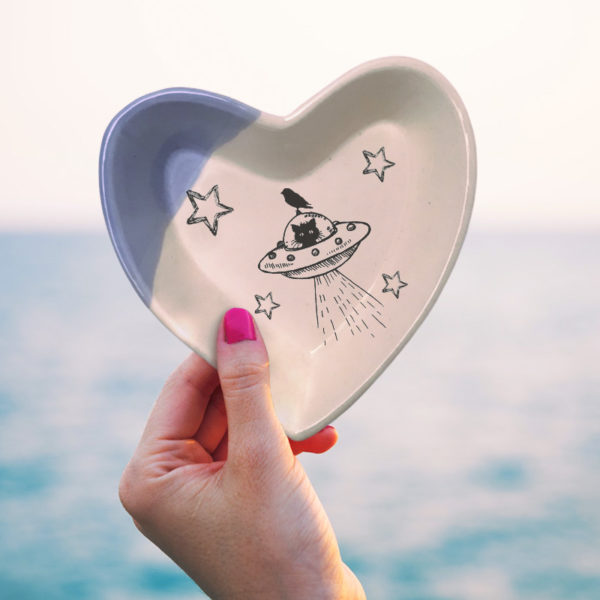 Handmade ceramic heart shaped dish with a drawing of a cat in a flying saucer or UFO. Lavender accent color.