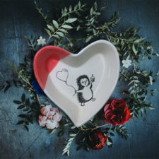 Handmade ceramic heart shaped dish with a drawing of hedgehog heading to a party with a cupcake and a balloon. Red accent color