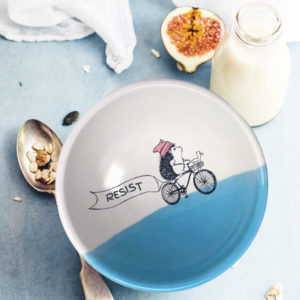 This handmade ceramic soup bowl has a drawing of a brave hedgehog resistance fighter, riding a bike with a huge resist banner. Blue accent color.