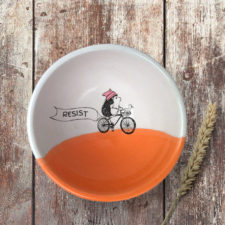This handmade ceramic soup bowl has a drawing of a brave hedgehog resistance fighter, riding a bike with a huge resist banner. Coral accent color.